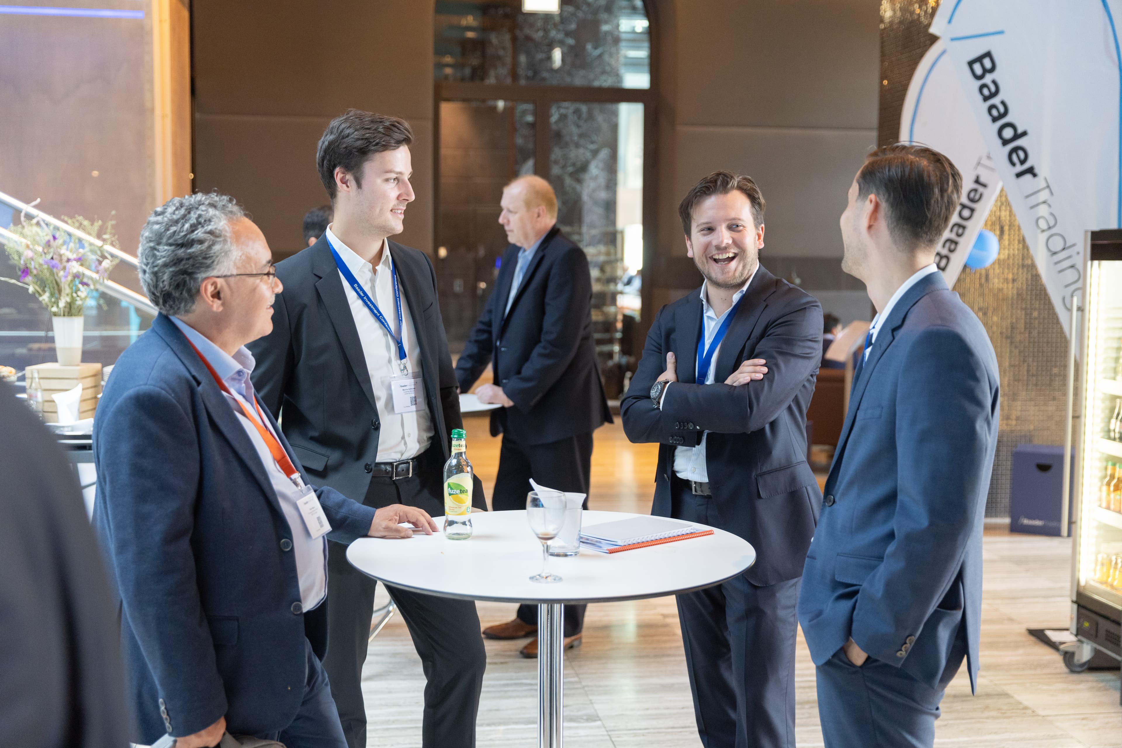 Vier Männer im Anzug stehen lachend an einem Stehtisch bei einer Baader-Bank-Veranstaltung.