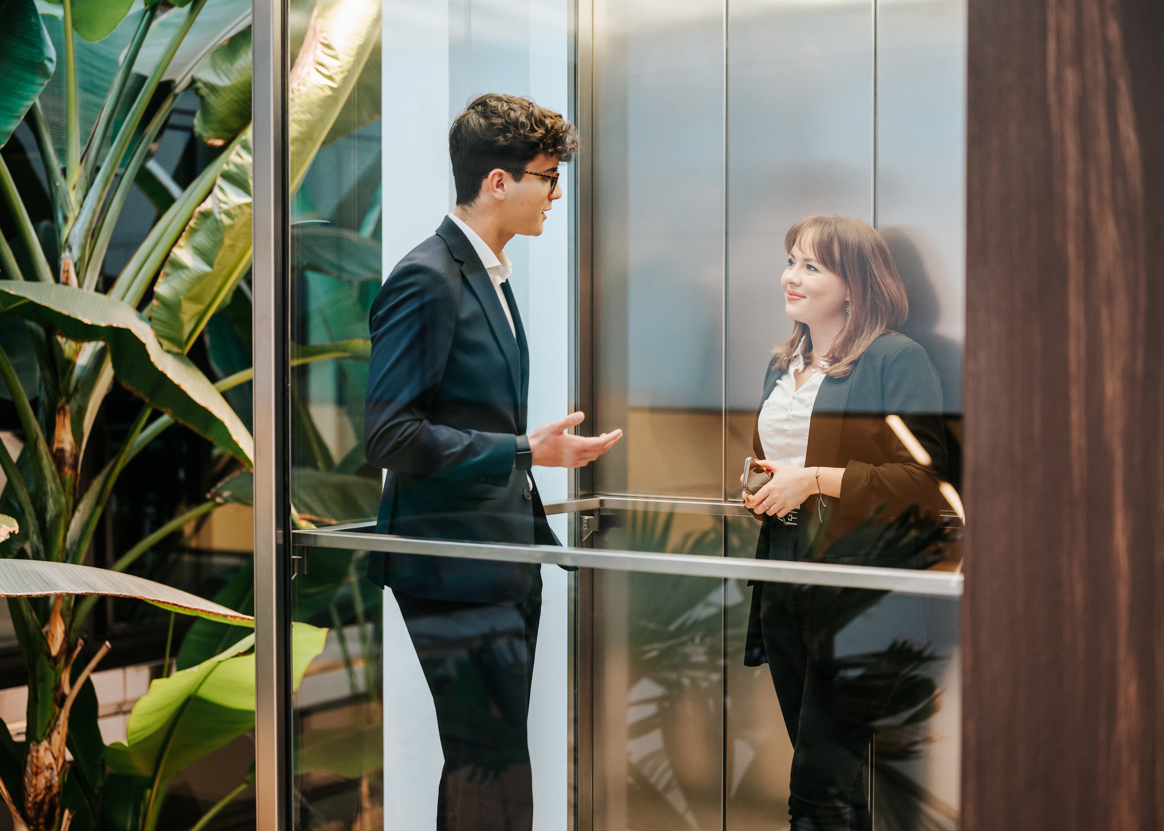 Zwei Baaderbank Mitarbeiter im Anzug im Aufzug, Gesprächsszene im modernen Atrium mit Pflanzen.