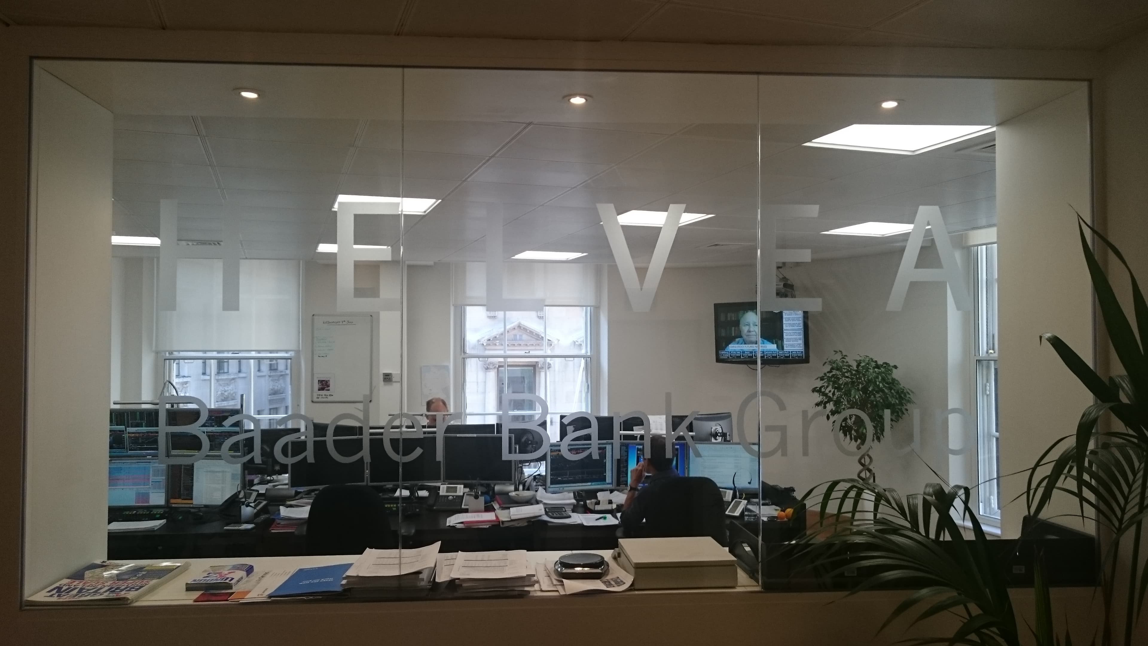Glaswand mit Aufschrift "Baader Bank Group" im Londoner Büro der Baaderbank, dahinter Arbeitsplätze.