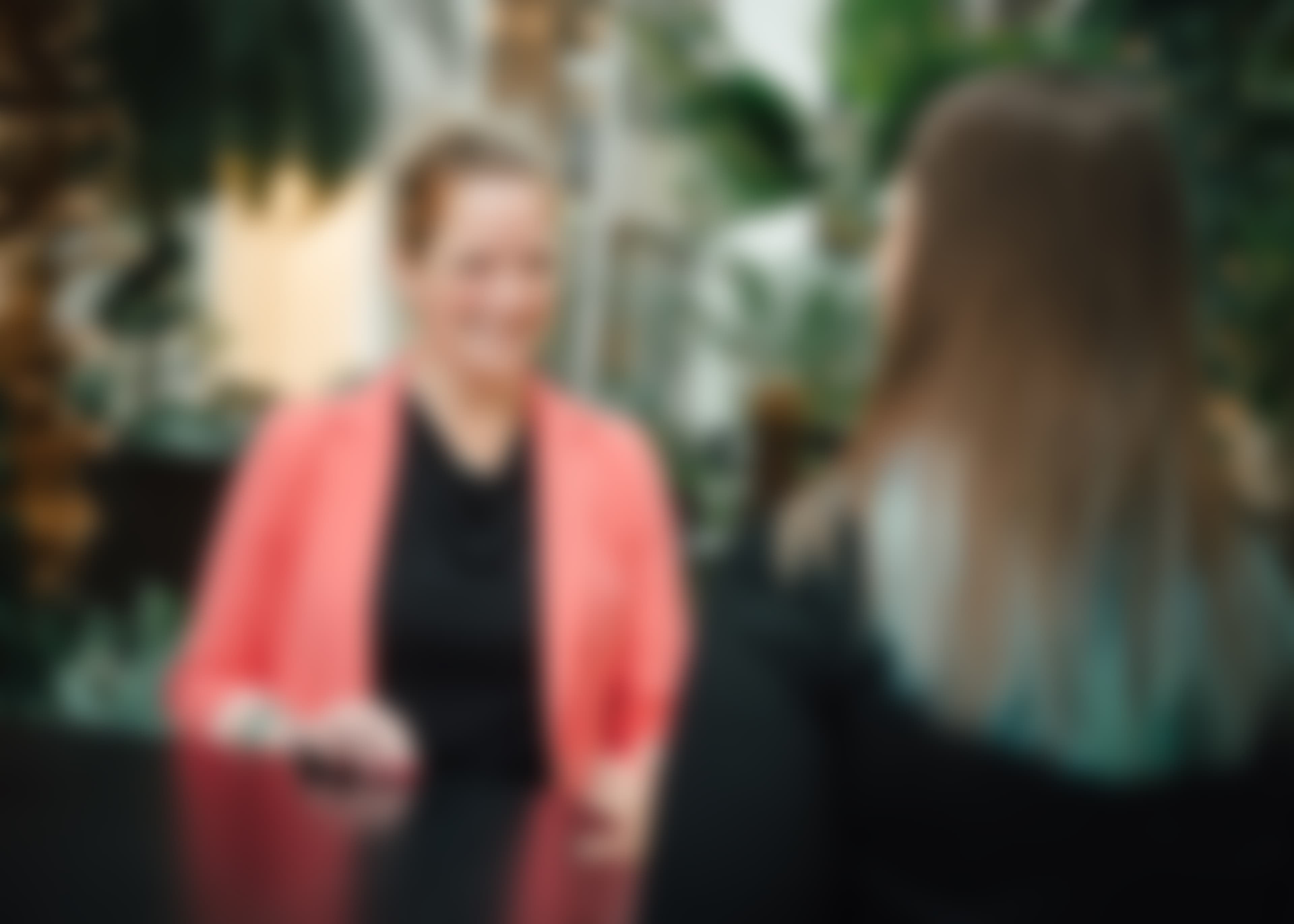 Unscharfe Aufnahme von zwei Frauen an einem Tisch, im Hintergrund sind große grüne Pflanzen erkennbar.