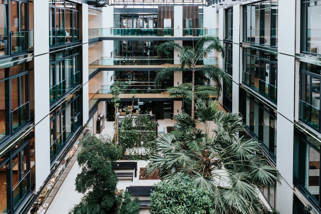 Baaderbank Atrium in Unterschleißheim mit moderner Glasfassade, tropischen Pflanzen und offenen Arbeitsbereichen.