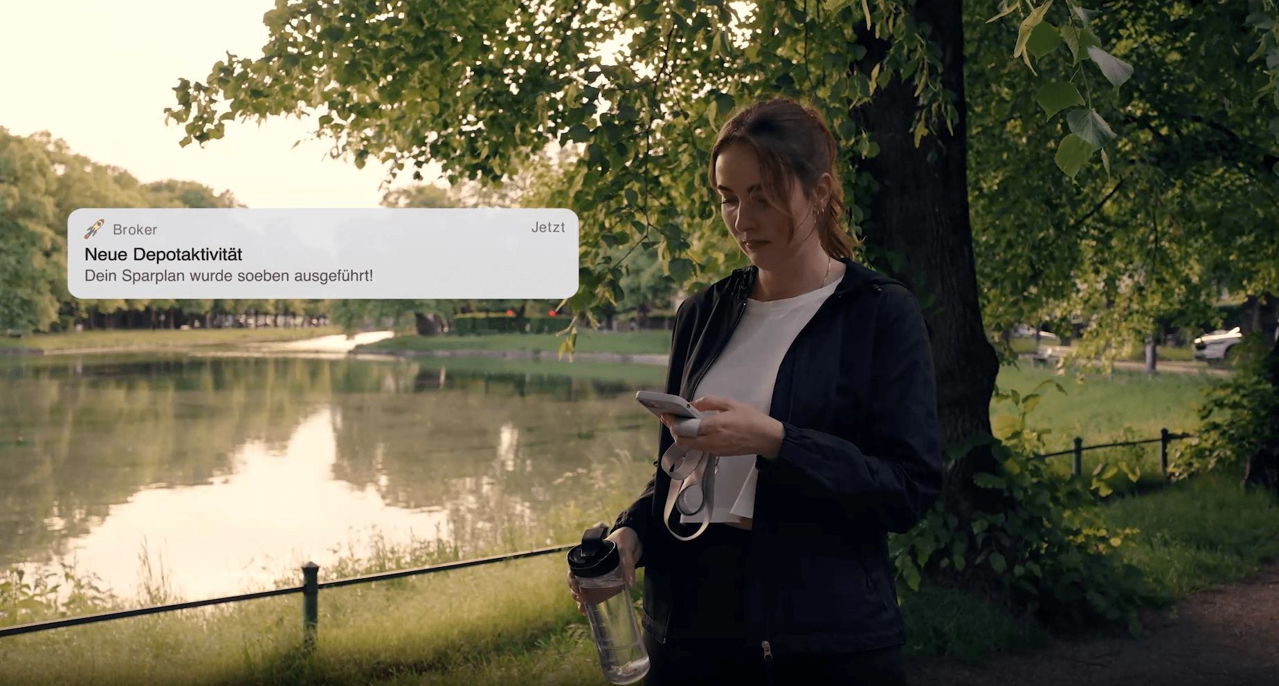 Eine Frau steht in Sportkleidung mit Trinkflasche und Smartphone in der Hand in einem Park. Auf ihrem Handy erscheint eine Push-Benachrichtigung mit dem Text „Neue Depotaktivität – Dein Sparplan wurde soeben ausgeführt!“.