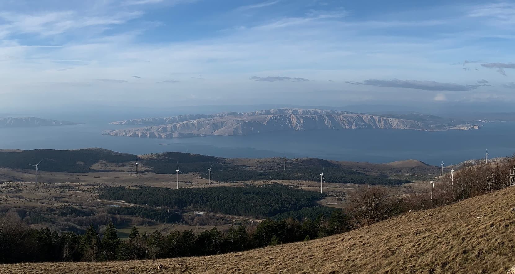 Landschaft mit mehreren Windrädern, dahinter Meer und felsige Inseln unter blauem Himmel.